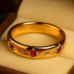 Puede incluir: Un anillo de oro con piedras preciosas rojas y pequeños diamantes, presentado en una caja de terciopelo marrón. El anillo presenta tres piedras rojas y pequeños diamantes engastados en una banda. El anillo es una joya.