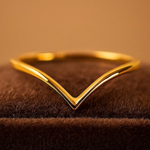 May include: A gold-colored ring with a V-shaped design. The ring is set on a brown velvet surface. The ring's design is simple and elegant, with a polished finish. The ring is a piece of jewelry.