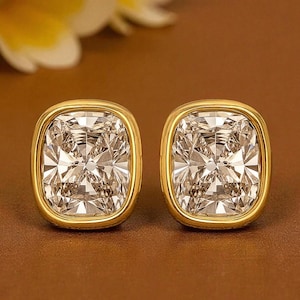 Peut inclure: Une paire de boucles d'oreilles carrées à clous dorées avec une grosse pierre claire et facettée. Les boucles d'oreilles ont une bordure dorée et sont posées sur un fond marron. Bijou classique.
