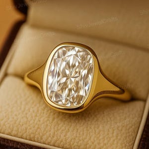 May include: A gold ring with a large, rectangular, clear gemstone set in a bezel setting. The ring is displayed in a brown jewelry box with a tan interior. The ring's band is smooth and polished.