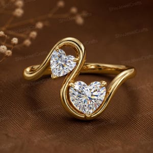 May include: A gold bypass ring featuring two heart-shaped clear stones set in a smooth, polished yellow gold band. The design showcases a flowing, asymmetrical silhouette that curves around each gemstone, creating a continuous, elegant look.