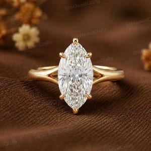 Puede incluir: Un anillo de oro con una gran piedra preciosa transparente de talla marquesa sujeta por seis garras. La banda tiene un diseño de vástago dividido donde los brazos de metal se encuentran con el engaste central, creando un aspecto pulido y liso.