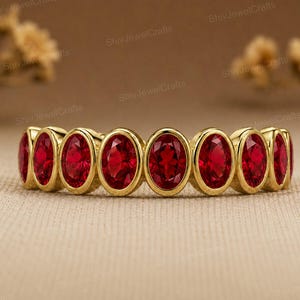 May include: A gold band ring featuring a series of oval-cut, deep red gemstones set in individual bezels. The ring displays a continuous row of these vibrant red stones, each framed by a polished gold metal setting, creating a uniform design.