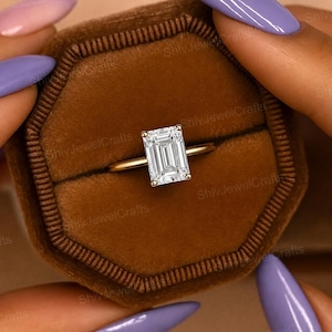Puede incluir: Un anillo de oro con una gran gema clara de forma rectangular. El anillo se presenta en una caja de terciopelo marrón. El anillo lo sostiene una persona con esmalte de uñas morado.