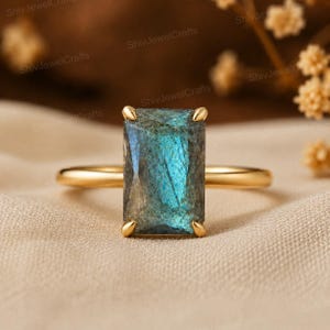 May include: A gold ring featuring a rectangular, faceted labradorite gemstone with a prominent blue iridescent flash. The stone is held in a four-prong setting on a thin, polished gold band, showcasing the natural color variations of the gem.