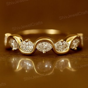 Könnte beinhalten: Ein goldfarbener Ring mit einer Reihe von ovalförmigen, klaren Edelsteinen, die in einem wellenförmigen Design gefasst sind. Der Ring hat eine polierte Oberfläche, die das Licht reflektiert. Die Edelsteine sind gleichmäßig über das Band verteilt. Ein Schmuckstück.