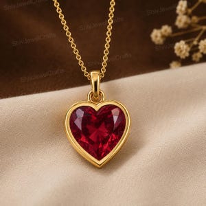 May include: A gold necklace featuring a heart-shaped pendant with a faceted deep red gemstone. The pendant is suspended from a delicate gold chain, showcasing a polished metal setting that frames the vibrant red heart-shaped stone clearly.