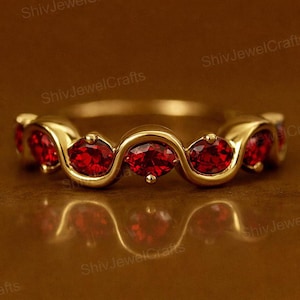 May include: A gold ring with a series of bright red gemstones. The oval-shaped gemstones are set in a wavy, gold band. The ring is displayed on a reflective surface, enhancing its visual appeal. The ring is a piece of jewellery.