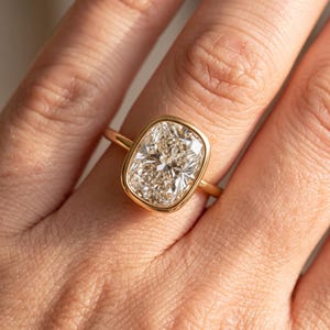 May include: A gold ring featuring a large, rectangular cushion-cut diamond set in a smooth, polished bezel setting. The band is thin and circular, crafted from yellow gold, providing a clean and classic look that highlights the center stone.
