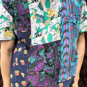 May include: A short-sleeved patchwork shirt featuring a mix of fabrics including a white panel with black line art of 1990s icons, a dark blue panel with a french fry and burger print, a purple geometric patterned panel, and a blue striped panel.