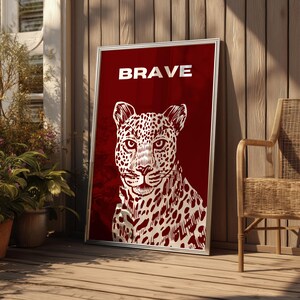 May include: A framed art print featuring a white leopard illustration on a deep red background. The word "BRAVE" is displayed in white at the top. The artwork is leaning against a wooden wall, next to a wicker chair.