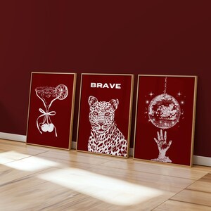 May include: Three framed art prints with a red background and gold frames. The prints feature white line drawings of a cocktail, a leopard with the word "BRAVE", and a disco ball. The prints are leaning against a red wall.