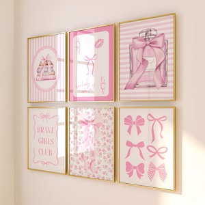 May include: Six framed art prints with gold frames. The prints feature pink and white designs, including bows, perfume, and the text "Brave Girls Club." The artwork has a feminine aesthetic.