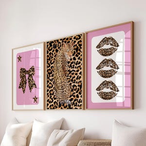 May include: Three framed art prints featuring a leopard print bow, a leopard, and three leopard print lips. The prints have pink and white backgrounds and are in gold frames. The leopard is wearing sunglasses.