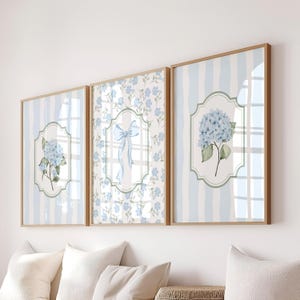 Könnte beinhalten: Drei gerahmte Wandkunstwerke mit Aquarellillustrationen von blauen Hortensien und einer blauen Schleife. Die Kunstwerke sind vor einem Hintergrund aus hellblauen und weißen Streifen und floralen Mustern angeordnet, alles in Holzrahmen.