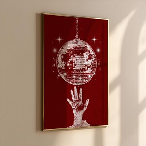 May include: A framed art print featuring a red background with a silver disco ball suspended from a chain. A hand reaches up towards the disco ball. The artwork is framed in a gold-colored frame.