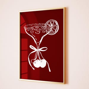 May include: A framed art print featuring a white line drawing of a margarita cocktail with a lime slice and cherries on a deep red background. The print is in a gold frame.