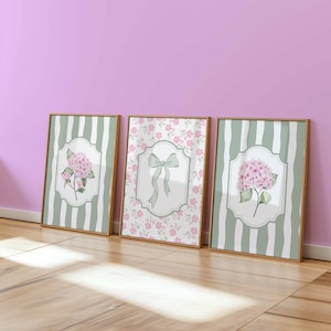 May include: Three framed art prints with floral and bow designs. Two prints feature pink hydrangea blooms on a white background with green striped borders. The center print showcases a green bow on a floral background. All are in gold frames.