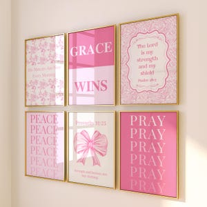 May include: A set of six framed wall art prints in shades of pink and white. The prints feature floral patterns, a pink bow, and religious text including Grace Wins, The Lord is my strength and my shield, Peace, Proverbs 31:25, and Pray.