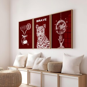 May include: Three framed art prints with a burgundy background and white illustrations. One print features a cocktail, another a leopard with the word "BRAVE", and the third a disco ball. The frames are a light wood color.