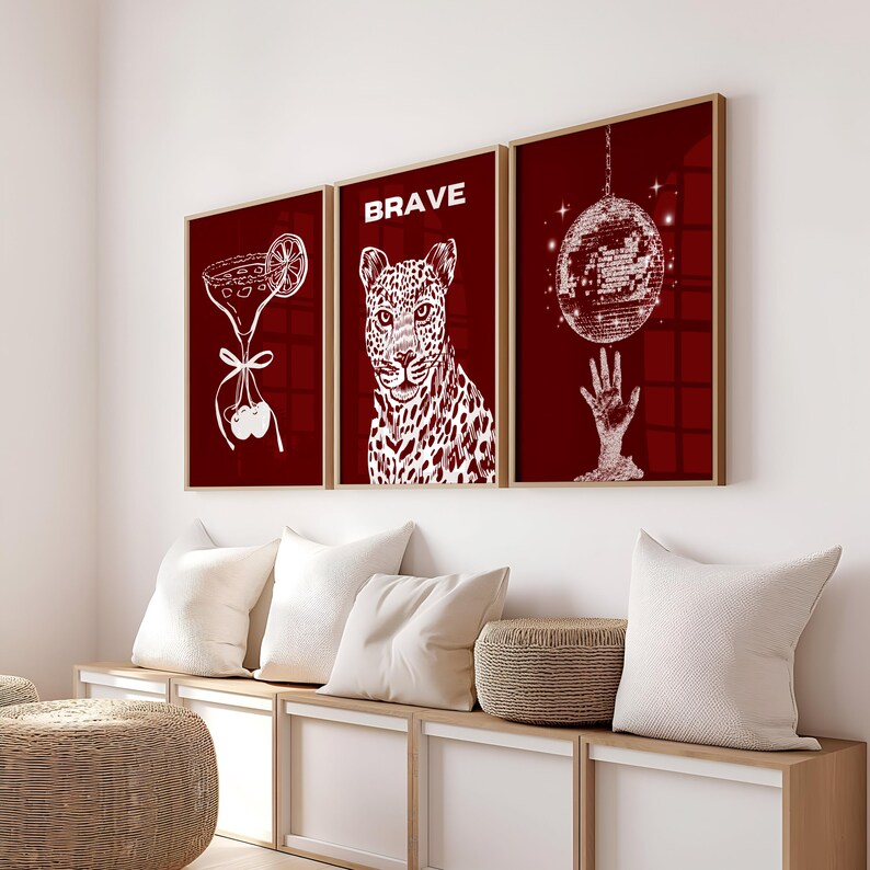 May include: Three framed art prints with a burgundy background and white illustrations. One print features a cocktail, another a leopard with the word "BRAVE", and the third a disco ball. The frames are a light wood color.