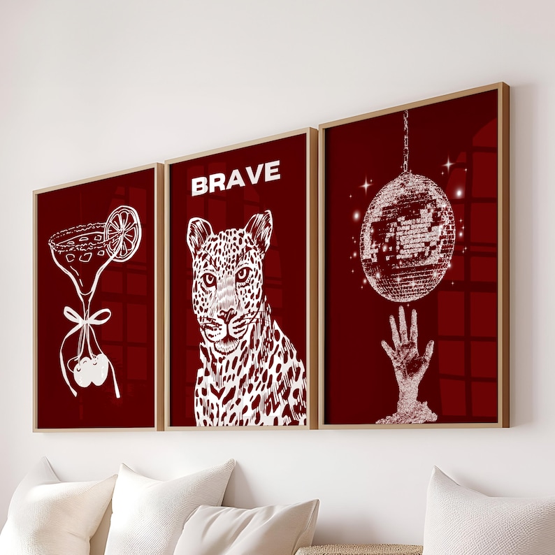 May include: Set of three framed art prints with a burgundy background. The prints feature white illustrations of a cocktail, a leopard with the word "BRAVE", and a disco ball above a hand. The frames are a light brown color.