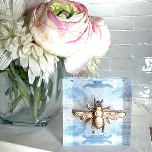 May include: A gold-colored bee sculpture is affixed to a square, light blue block with a cloudy design. The bee has detailed wings and body. A glass vase with white and pink flowers is in the background.