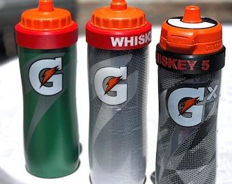 May include: Three Gatorade squeeze bottles with orange caps. The first is green with a white G logo. The second is grey with a white G logo and the text WHISKEY. The third is clear with a black grid pattern, a Gx logo, and the text WHISKEY 5.