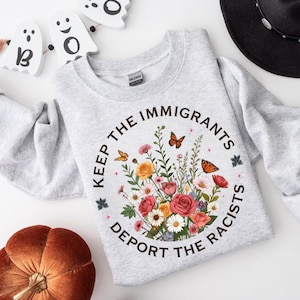 May include: A light gray sweatshirt with the text "KEEP THE IMMIGRANTS DEPORT THE RACISTS" in a circular design. The design includes a floral arrangement with butterflies. A black hat, a pumpkin, and Halloween decorations are in the background.
