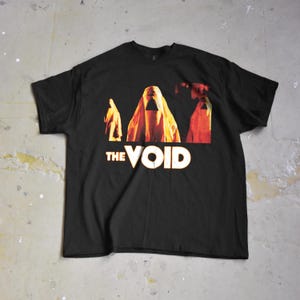 Op de afbeelding: Zwart t-shirt met een afbeelding van drie figuren in oranje en gele gewaden, met de tekst "THE VOID" in wit en goud. Het ontwerp is gecentreerd op de voorkant van het shirt.