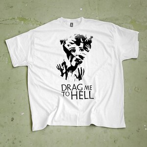 Pode incluir: Camiseta branca com um gráfico preto de um rosto angustiado com mãos e o texto "DRAG ME TO HELL". A camiseta tem gola redonda e mangas curtas. O design está centralizado na frente.