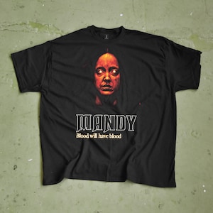 May include: Black t-shirt featuring a graphic from the movie "Mandy." The design includes a portrait of a person with glowing eyes, the title "MANDY," and the phrase "Blood will have blood" in white text. The shirt is on a green surface.