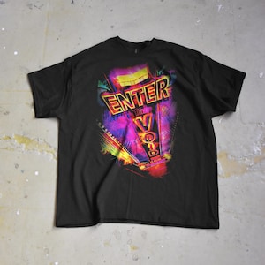 Puede incluir: Camiseta negra con un diseño gráfico vibrante. El diseño presenta las palabras "ENTER THE VOID" en una fuente de estilo neón, con un fondo abstracto colorido en tonos morado, rosa, amarillo y verde. La camiseta está tendida sobre una superficie de hormigón.