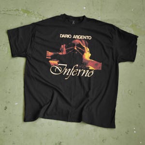 Może przedstawiać: Czarny t-shirt z napisem "DARIO ARGENTO" nad grafiką z filmu "Inferno". Grafika przedstawia osobę zasłaniającą twarz dłońmi, z czerwonymi i żółtymi akcentami. Widnieje również słowo "Inferno".