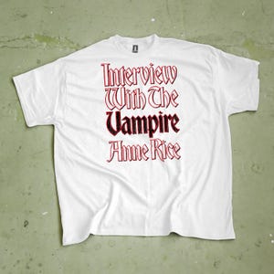 Könnte beinhalten: Weißes T-Shirt mit dem Text "Interview With The Vampire Anne Rice" in roter und schwarzer gotischer Schrift. Das Shirt besteht aus einem weichen Material und liegt flach auf einer grünen Oberfläche.