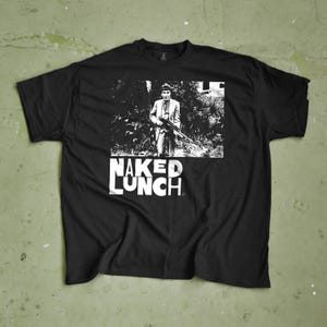 May include: Black t-shirt featuring a black and white image of a person holding a rifle, with the text "NAKED LUNCH" in white block letters below the image. The shirt is made of a soft material.