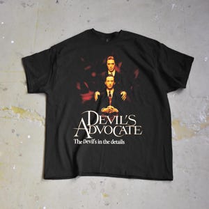 May include: Black t-shirt featuring a movie poster design. The graphic includes two men in suits, with the words "Devil's Advocate" and "The Devil's in the details" in white text. The shirt is laid flat.