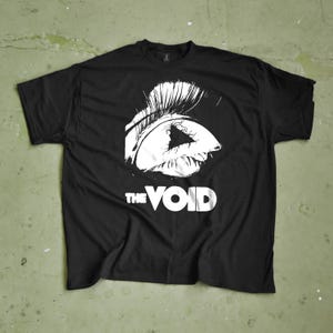 May include: Black t-shirt featuring a white graphic of an eye and the text "THE VOID". The eye design is detailed with white lines and shading, contrasting against the black fabric. The shirt is displayed flat, showcasing the design. The font is bold.