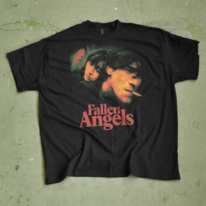 May include: Black t-shirt with a graphic of two people and the text "Fallen Angels" in red. The graphic has a vintage, faded appearance. The shirt is laid flat on a green surface.