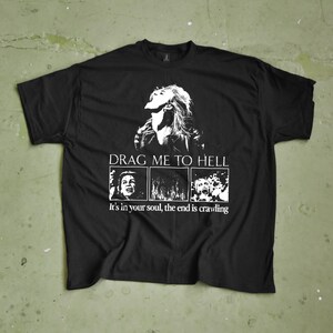 Pode incluir: Camiseta preta com um design gráfico. O design apresenta o rosto de uma mulher e o texto "DRAG ME TO HELL". Abaixo, três painéis com imagens e a frase "It's in your soul, the end is crawling."