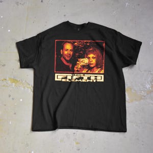 Op de afbeelding: Zwart T-shirt met een filmposter-afbeelding van "The Fifth Element". De afbeelding toont twee mensen en de titel van de film in een vetgedrukte, gestileerde lettertype. Het shirt is gemaakt van een zacht materiaal.