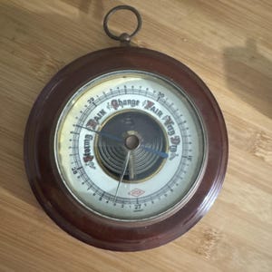 May include: A vintage barometer with a dark brown wooden frame and a white face. The face features a needle pointing to the current atmospheric pressure, with text indicating "Stormy", "Change", and "Fair". A metal ring is at the top.