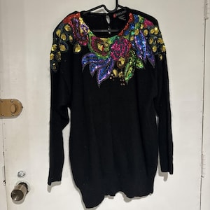 May include: Black sweater with a round neckline and long sleeves. The sweater is adorned with a colorful sequined floral design across the shoulders and chest. The sequins are in shades of gold, red, blue, and green.