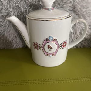 May include: White ceramic teapot with a lid and handle. The teapot features a pink and gold emblem with a bird illustration. The lid and emblem have pink and blue accents. The spout and handle are white with gold trim.