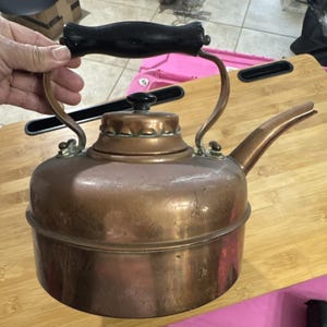 May include: A copper-colored kettle with a black handle and spout. The kettle has a round body and a lid with a small black knob. The handle is arched and attached to the body with brass fittings. A vintage kitchenware item.