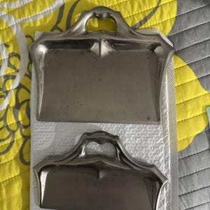 May include: Two antique silver-toned dustpans with heart-shaped handles. The dustpans have a rectangular shape with a slightly curved edge. The handles are arched and feature a decorative design. The dustpans are placed on a white surface.