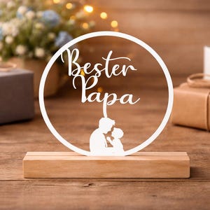 Puede incluir: Un letrero de madera blanco con las palabras "Bester Papa" en un diseño circular, con una silueta de un padre y un niño. El letrero está sobre una base de madera, con regalos y flores en el fondo. Tema: regalo del Día del Padre.