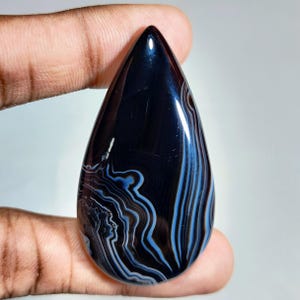 May include: A polished, teardrop-shaped gemstone with a dark blue base and intricate banding in shades of white, brown, and light blue. The stone is held between fingers, showcasing its smooth, reflective surface. The gemstone is a natural stone.