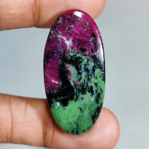 May include: An oval-shaped gemstone with a vibrant mix of colors. The top half is a deep magenta, transitioning to black and then a speckled green at the bottom. The stone is held between two fingers.