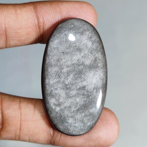 Puede incluir: Gema ovalada pulida con un brillo plateado. La piedra tiene una superficie lisa y reflectante con un patrón sutil y texturizado. La gema se sostiene entre los dedos, mostrando su tamaño y forma.